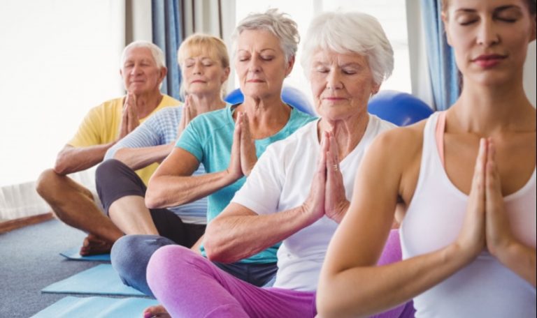 A group of seniors sit on mats doing yoga with hands in prayer pose, eyes closed, and looking calm.