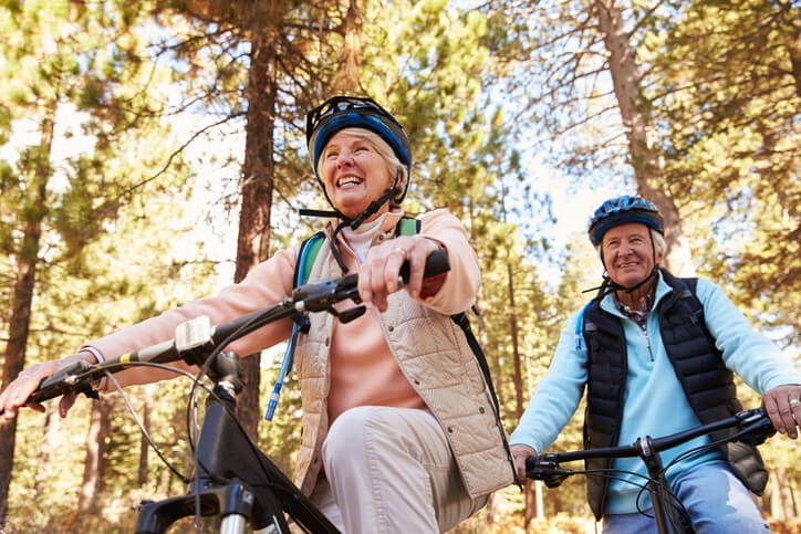 Two smiling older adults ride bicycles together through a sunlit forest, wearing helmets and casual outdoor clothes.